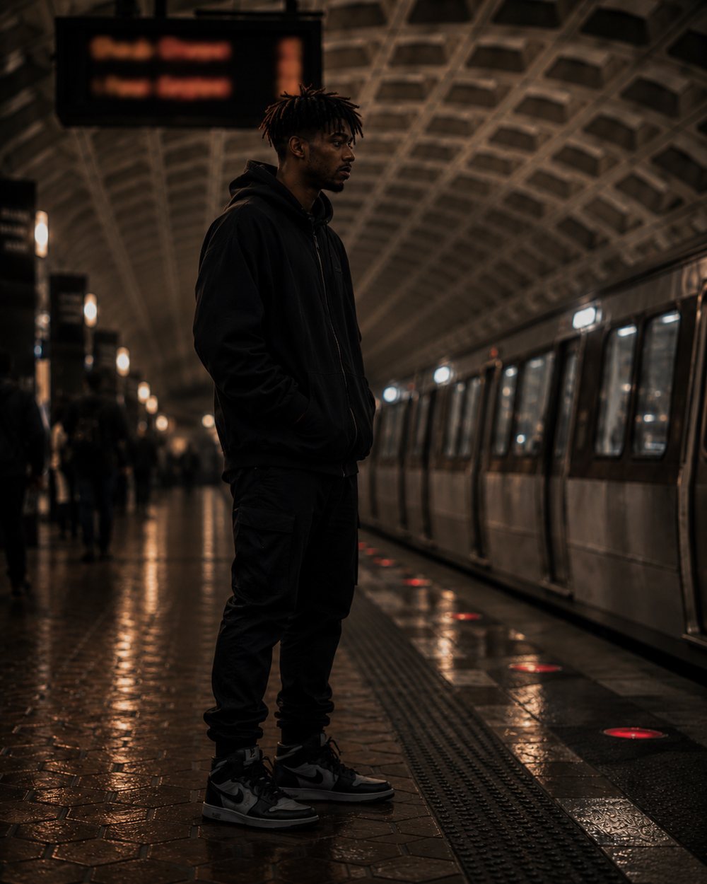 Kwame in a black hoodie and sneakers standing on a Metro platform beside a waiting train